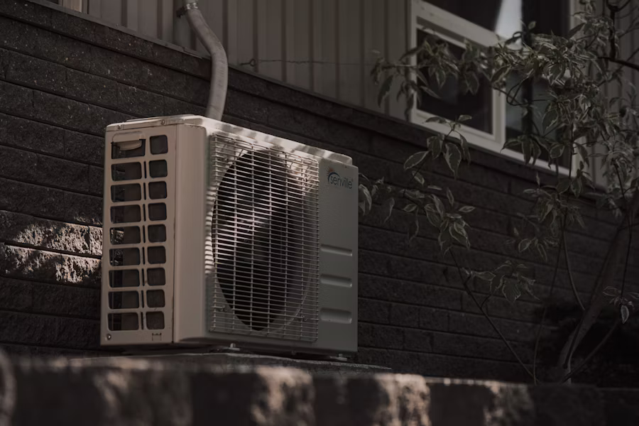 Residential air-conditioning condenser unit on the side of a suburban home in late afternoon light, framed by foliage and the home's siding.